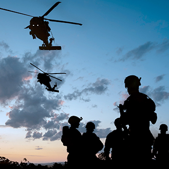 Silhouettes of five army men in the field at dusk with two helicopters flying above