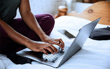 Close-up of a person sitting cross-legged on a bed typing on a laptop